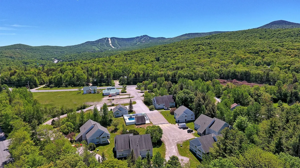 A wide-angle shot showing wooded mountains, scattered houses, green open spaces, and the swimming pool. The composition highlights the connection between the resort buildings and the surrounding natural landscape, showcasing both the man-made amenities and the wild Vermont outdoors.
