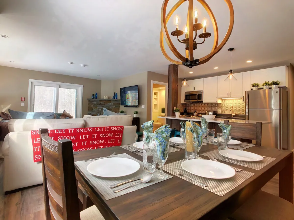 The image showcases a warmly lit open-concept living and dining area. A rustic-modern dining table, elegantly set for a meal, sits beneath a stylish chandelier. Nearby, a plush sofa adorned with decorative pillows invites relaxation, while the overall ambiance is cozy and perfect for gatherings.
