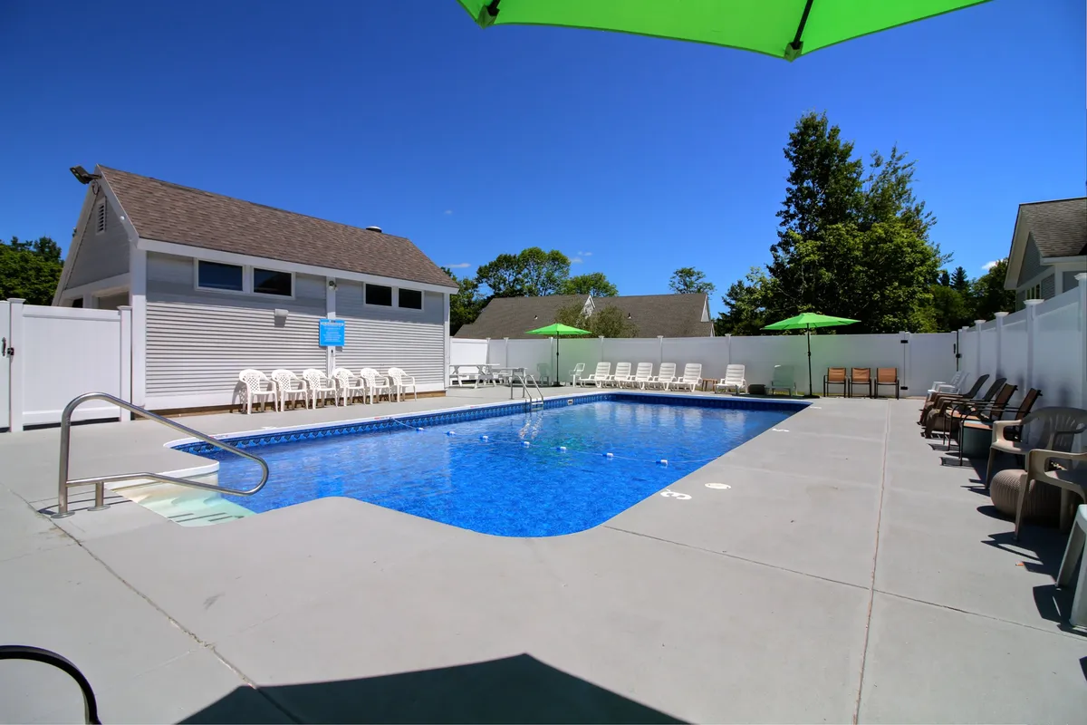 A vibrant outdoor swimming pool surrounded by lounge chairs and green umbrellas, with lush greenery framing the edges. The setting exudes a cheerful, retreat-like atmosphere ideal for summer relaxation and poolside gatherings.