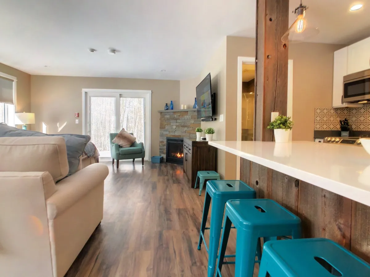 This image captures a bright, open-concept living space featuring a cozy sofa adjacent to a gas fireplace, a TV, a kitchen island with seating, and a green accent chair. Natural light floods the room, highlighting the blend of modern and rustic elements.