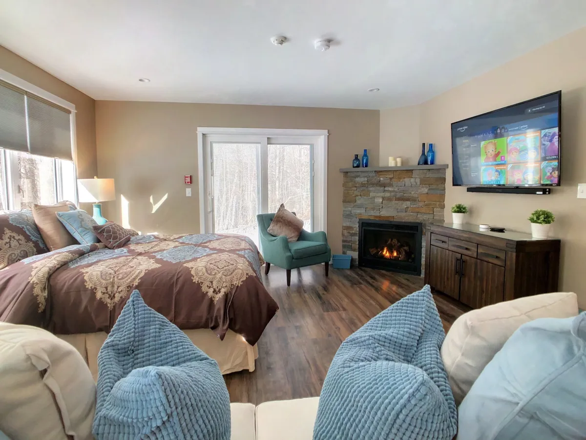 The scene features a bed with decorative bedding, a glowing fireplace, a television, and plush cushions. The inviting setup suggests cozy nights spent relaxing by the fire, watching movies, or enjoying peaceful mornings together.