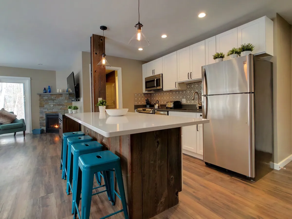 This image provides a wide-angle view of the open-plan space, including a kitchen island with bar stools, stainless appliances, and a stone fireplace, all illuminated by warm lighting. It captures both the kitchen and living area in one shot.