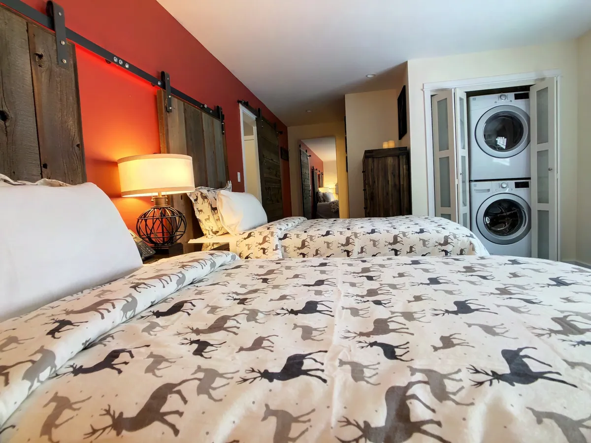 A cozy interior view highlighting modern washer and dryer units next to a bed with reindeer-patterned bedding and wooden sliding doors. The mood is warm and inviting, suggesting comfort and practicality for longer stays.