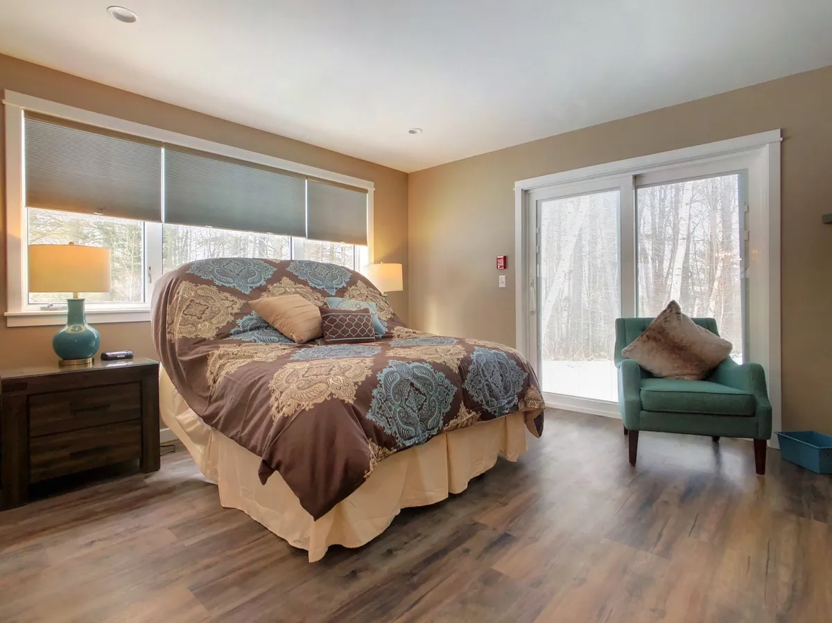 A spacious and inviting master bedroom featuring a large bed with decorative bedding, a cozy armchair, and expansive windows that let in abundant natural light. The setup emphasizes both relaxation and flexibility, as the space appears suitable for couples or can be adapted for different sleeping arrangements.