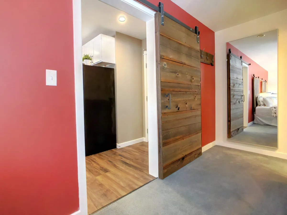 The image captures a welcoming interior scene featuring sliding barn doors, a striking bright red accent wall, and a mirror reflecting the room’s cozy ambiance. The setting suggests moments of quiet relaxation or lighthearted conversation near the entrance, with farmhouse-chic décor touches evident throughout.