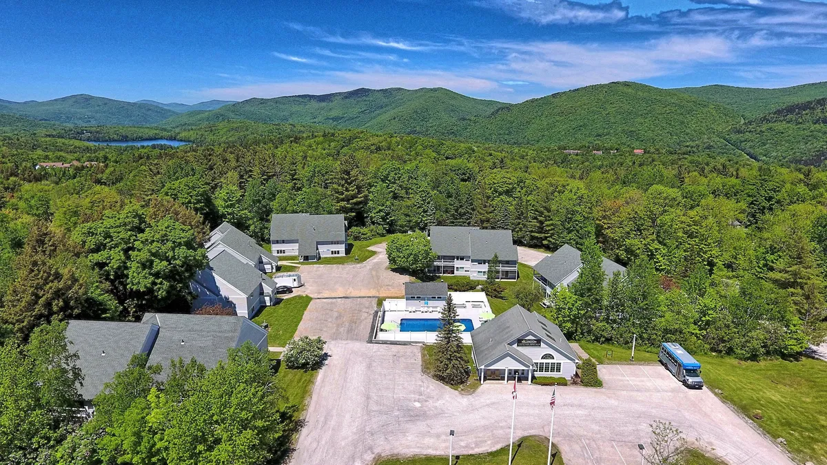 A scenic view of the property’s exterior, featuring the swimming pool surrounded by lounge chairs and lush trees, with mountains rising in the background and several buildings nestled into the landscape. The pool is the centerpiece, while the expansive mountain vista beckons adventure.