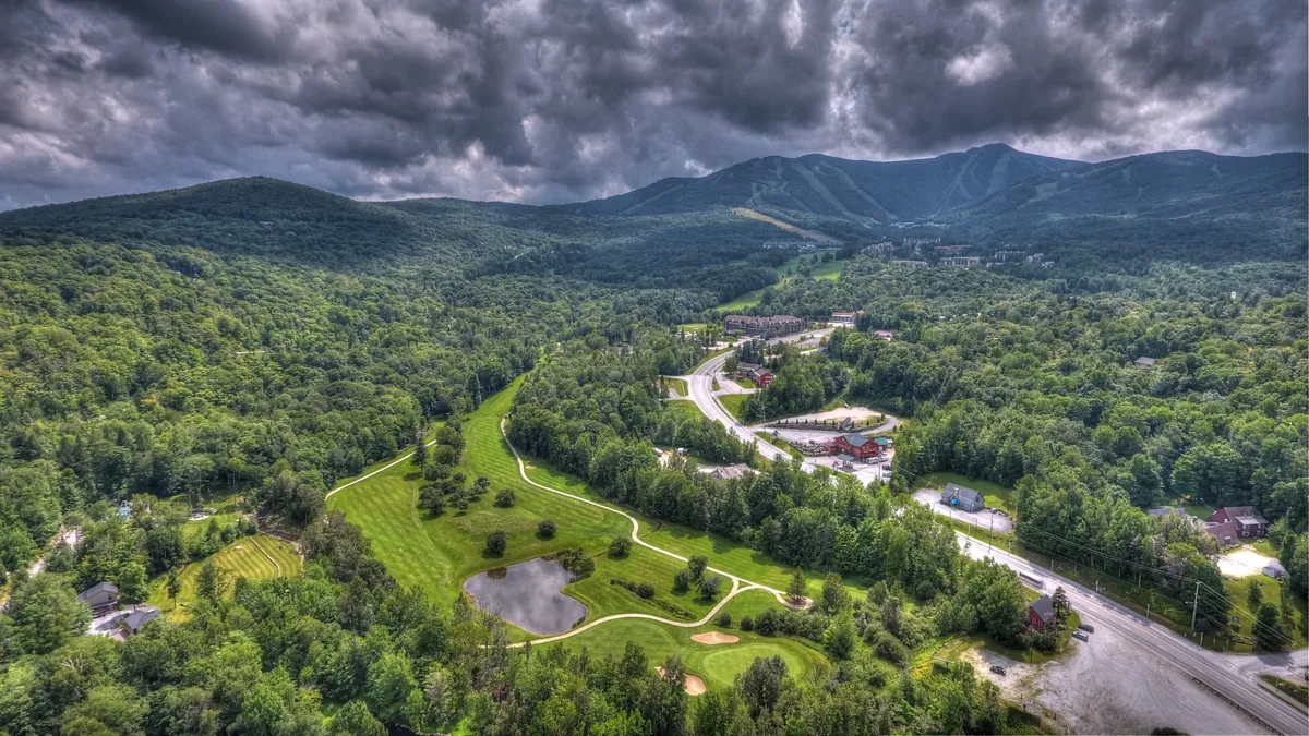 A wide view of Vermont’s landscape featuring a mountain range, a well-manicured golf course, winding road, pond, and dense forests. The scene exudes tranquility and offers a sense of the vast array of outdoor activities available, including hiking, golfing, and nature walks.