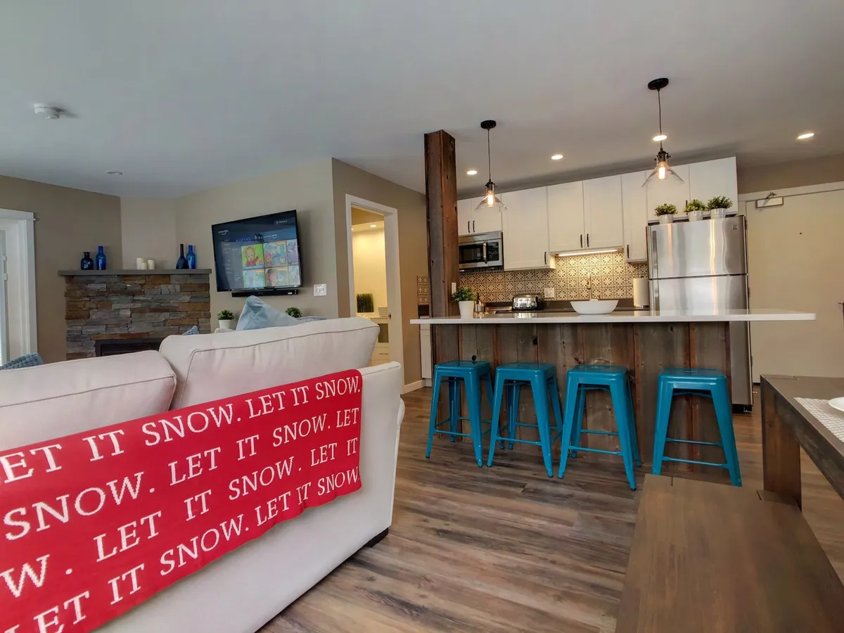 A view of a stylish kitchen island with stools, a stone fireplace, and a cozy living space with a red blanket. The scene captures a social, communal vibe, ideal for entertaining friends or enjoying late-night snacks after exploring local venues.